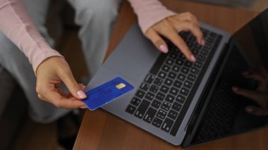 Oturma odasında dizüstü bilgisayar kullanan bir kadın, elinde modern alışverişi gösteren rahat bir ev içinde tasvir edilmiş kredi kartı tutuyor.