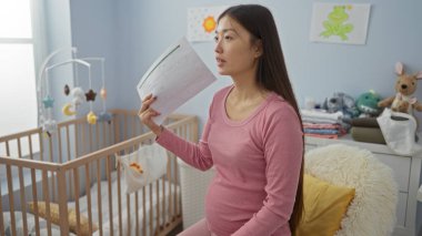 Hamile kadın sıcak bebek odasında elinde beşiği ve oyuncakları olan bir belge tutuyor, müstakbel annelik anlarını yakalıyor ve ev yaşamını ve hazırlıklarını özendiriyor..