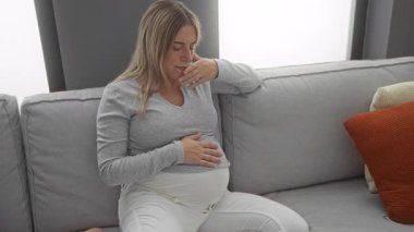 Oturma odasında kanepede oturan hamile bir kadın rahat bir dairede el üstünde endişeli endişeli bakıyor.