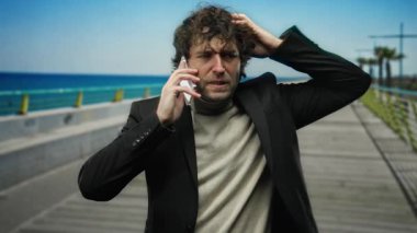 Stresli genç İspanyol bir adam deniz kenarında telefonla konuşuyor, şık siyah bir ceketle, kafası karışmış bir ifadeyle, dışarıda manzaralı bir plaj zeminine karşı duruyor..