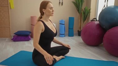Spor salonundaki mavi bir paspasın üzerinde yoga yapan bir kadın. Etrafı spor aletleriyle çevrili. Toplar, bitkiler, somut odak ve sükunet de dahil..