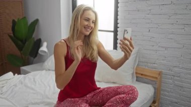 Yatakta oturan bir kadın, parlak yatak odasında selfie çekiyor. Yanında bitkiler olan kırmızı pijamalar giyiyor. Beyaz tuğlalı arka plan, rahat bir ev hayatı sergiliyor. 