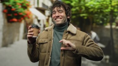 Şehrin dışında elinde kahve fincanı olan genç bir adam canlı, renkli bir caddede gülümseyip kamerayı işaret ediyor..