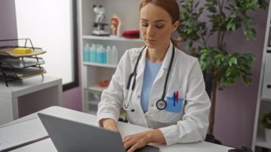 Klinik bir ortamda dizüstü bilgisayarda çalışan kadın doktor. Parlak, düzenli bir ofis alanında tıbbi ekipmanlarla çevrili. Bu da profesyonel bir sağlık hizmetleri ortamı olduğunu gösteriyor..