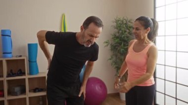 Orta yaşlı İspanyol bir çift spor salonunda bir kadınla birlikte acı çeken bir adama yardım ediyorlar. Etrafı egzersiz ekipmanlarıyla çevrili, canlı bir iç mekanda sağlıklarını ve desteklerini gösteriyorlar..