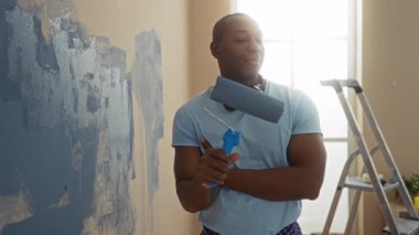 Oturma odasında duvarları boyayan, kulaklık takan, gülümseyen ev yenileme projesinin keyfini çıkaran genç bir adam..