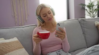 Evde dinlenen, elinde telefon ve fincanla oturma odasında kulaklık takan bir kadın..