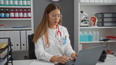 Gözlüklerini çıkaran, hastane ofisinde oturan, steteskoplu beyaz önlüklü bir kadın, masadaki tıbbi malzeme ve belgelerin arasında dizüstü bilgisayarla meşgul olan bir kadın..