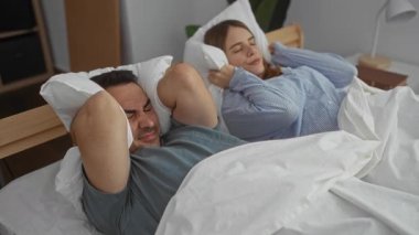 Kadın ve erkek yatakta kulaklarını kapatıyor, hayal kırıklığına uğramış, rahat bir yatak odası ortamında, bir çiftin evde gürültülü bir olay yaşadığını gösteriyor..
