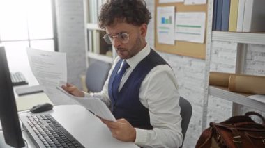 Ofiste çelik yelek ve gözlüklü bir adam, bilgisayar ve kitaplık ile masadaki belgelere odaklanmış, ciddi bir çalışma ortamı yaratıyor..