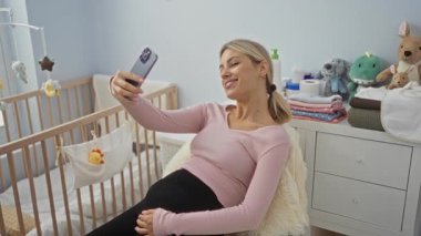 Yatak odasında beşiğin yanında selfie çeken, gülümseyen ve gevşemiş, sevimli oyuncaklar ve katlanmış giysilerle çevrili, sıcak ev ortamının tadını çıkaran bir kadın..