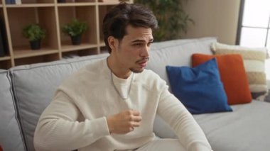 Sıcak bir oturma odasında, rahat görünümlü ve yelpaze giyen, renkli yastıklarla çevrili gündelik bir kazak giyen, kapalı alanda rahatlık ve rahatlama tasvir eden genç İspanyol bir adam..