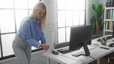 Modern ofis ortamında çalışan kadın, bilgisayar ve pencereleri doğal ışık ayarlarını vurgulayan belgeleri masanın yanında tutuyor.