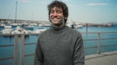 Deniz kıyısındaki bir limanda gülümseyen genç adam kamerayı işaret ediyor, açık bir günde bir tekne zeminine karşı duruyor..