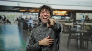 Kapalı bir kafede neşeli bir ifadeyle kameraya bakıp gülen genç bir adam, gündelik bir ortamda neşeli bir anı yakalıyor..