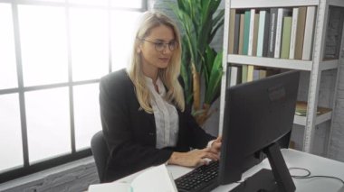 Ofiste çalışan bir kadın, deftere yazarken bilgisayar kullanıyor, etrafı kitaplarla ve yeşil bitkilerle çevrili, gözlük takıyor.. 