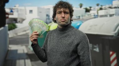 Geri dönüşüm konteynırının yanında Rus rublesi tutan İspanyol bir adam. Etrafı çöp ve kentsel ortamlarla çevrili. Paraya odaklanırken çeşitli duygular ifade ediyor..