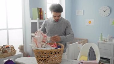 Çamaşırları parlak, düzenli bir çocuk odasında katlayan genç bir adam bir sepet giysi, yumuşak oyuncaklar ve ütü masasında ev bakıcılığı görevlerini vurgulayan bir ütü..