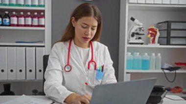 Üniformalı genç beyaz kadın doktor klinikte kol saatini kontrol ediyor, dakiklik ve profesyonelliğe odaklanmış.