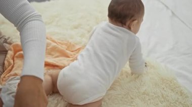 Anne sıcak bir yatak odası halısında bebeğini nazikçe değiştiriyor. Huzurlu bir anda sevgi ve aile bağını gösteriyor..