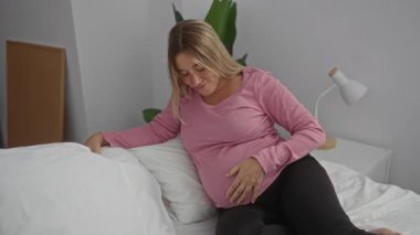 Sarışın hamile bir kadın rahat bir yatak odasında bir yatağa oturur, karnına hafifçe dokunur, evde sakin ve beklenen bir atmosferi canlandırır..