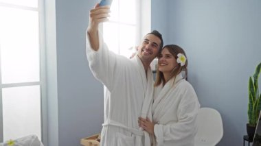 Kaplıca elbiseli bir çift evde selfie çekiyorlar, modern bir sağlık ortamında sevgi ve rahatlığı somutlaştırıyorlar..