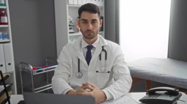 Klinikteki genç adam masasında steteskop ve beyaz önlükle oturuyor. Hastane odasında profesyonel ve yakışıklı görünüyor.