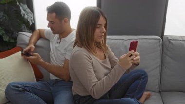 İspanyol çift rahat bir oturma odasında koltukta otururken cep telefonu kullanıyor, modern ilişki dinamiklerini ve dijital etkileşimi vurguluyorlar..