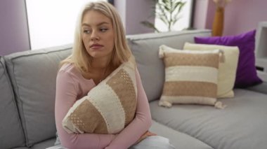 Evinde oturan kadın yastığını tutuyor. Rahat bir apartman dairesinde, yumuşak ışıklandırmalı ve modern dekorlu düşünceli görünüyor..