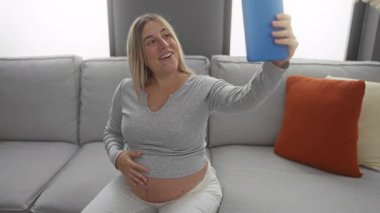 Sıcak bir oturma odasında kanepede oturup tabletindeki bir video çağrısını kaydederken gülümseyen ve kucağında bebek şişliği olan hamile bir kadın..