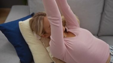 Rahat bir oturma odasında, kanepede uzanan, pembe bir bluz giyen, sarı saçlı, rahat bir şekilde gülümseyen, sessiz bir anın tadını çıkaran bir kadın..