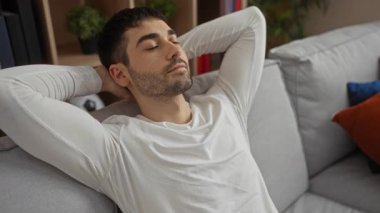 Rahat bir oturma odasındaki kanepede uzanan genç adam, gözleri kapalı ve elleri başının arkasında, modern bir ev ortamında huzur ve sükunet duygusu taşıyor..