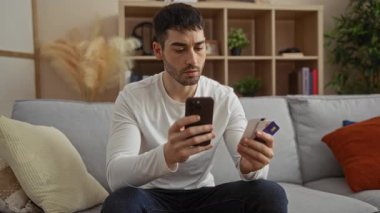 Evde elinde akıllı telefon ve kredi kartı olan genç adam oturma odasında konsantre olmuş mali planlama ya da online alışveriş önerileri sunuyor.