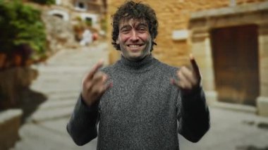 Genç bir adam şehir ortamında taş gibi bir jest yapıyor, rahat bir şekilde gülümsüyor şehir arka planı ve şık tavırlarıyla, canlı sokak havaları sergiliyor..