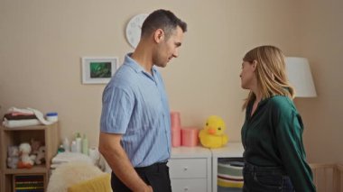 Konforlu bir yatak odasında duran bir çift, görünür bir beşik ile konuşuyor, aile hayatına ve ilişkilere odaklanmış huzurlu bir ev ortamını öneriyor..