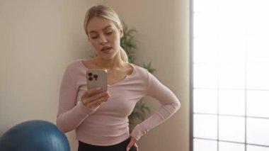 Spor merkezinde spor topu ve direniş bantlarıyla akıllı telefon kullanan bir kadın, spor yaparken modern fitness ve teknoloji bağlantısı sergiliyor..
