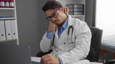 Klinikte çalışan, gözlük takan ve steteskop kullanan genç bir adam laptopa dikkatle odaklanıyor..