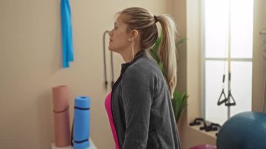 Parlak bir dekorla spor salonunda egzersiz yapan, pembe bluzlu ve gri ceketli, havluları tutan, spor aletleri ve yoga minderleriyle çevrili, evden hoşlanan bir kadın..