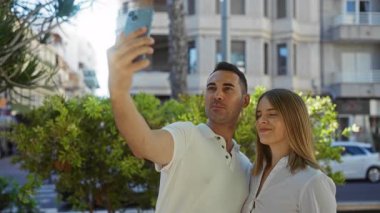 Güneşli bir sokakta gülümseyen ve selfie çeken bir çift. Şehir arka planında mutlu anları yakalıyorlar.