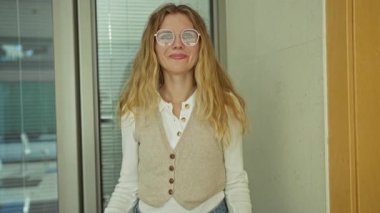 Ofiste gülümseyen, iş ortamında olumlu duygular ifade eden, gözlüklü, sarı saçlı, sıradan iş kıyafetleri giyen bir kadın..