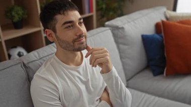 Modern oturma odasında dinlenen genç bir adam. Düşünceli bir yüz ifadesi var. Rahat gri bir koltuğa oturmuş, şık bir iç mekan.