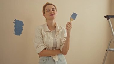 Oturma odasının duvarlarını boyayan bir kadın elinde fırça, yakınındaki merdivenle güvenle gülümseyen bir kadın ve kapalı arka planda yeni boyanmış bir duvar. 