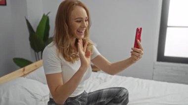 Parlak bir yatak odasında, yumuşak dekorasyon ve doğal ışıkla çevrili, gülümseyen ve el sallayan bir kadının akıllı bir telefonda görüntüsü..