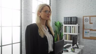 Gözlüklü, sarı saçlı, kendine güvenen genç bir kadın, ve modern ofis ortamında, bitkiler, raflar ve bilgisayarlarla çevrili, profesyonel bir atmosferi yansıtan,. 