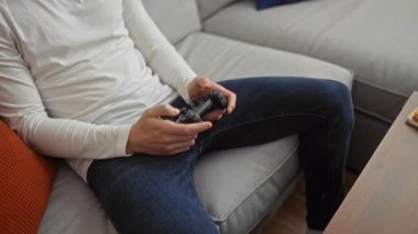 Modern bir apartman dairesinde rahat bir koltukta oturan bir adam video oyunları oynuyor. Etrafında samimi bir ifade ve rahat bir ortam yaratıyor.