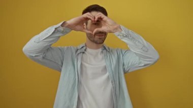 Gündelik kıyafetler giyen bir adam elleri ile kalp şekli oluşturan canlı sarı bir arka plana gülümseyen neşeli ve neşeli bir tavırla mutluluk ve sevgiyi ifade eden bir adam..