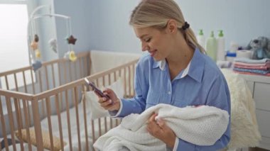Beyaz bir battaniyenin içinde bebeği tutarken gülümseyen bir kadın, yanında ahşap bir beşiği olan rahat bir yatak odasında, parlak ve huzurlu bir evde akıllı telefon kullanan bir kadın..