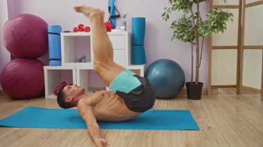 Spor salonunda yoga minderinde egzersiz yapan genç adam, spor aletleri ve aydınlık alanda ileri germe hareketleri yapan, atletik beceri gösteren ve sağlığa odaklanan biri..