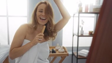Havluya sarılı, spa odasında tarakla şarkı söyleyen bir kadın. Neşeli bir ifadeyle ve parlak iç organlarıyla rahatlama ve sağlık ortamında mutluluk öneriyor.