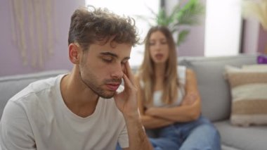 Kadın ve erkek, sıcak bir oturma odasında ciddi bir şekilde tartışıyorlar, bir ilişkide gerginliği vurguluyorlar, erkek gözle görülür şekilde stresli ve kadın arka planda oturuyor..